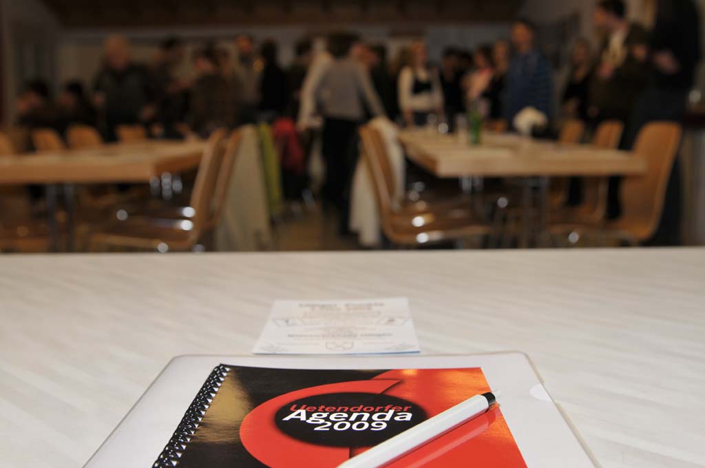 Eine Agenda von 2009 liegt auf einem Tisch. Weit im Hintergrund sind stehende Personen zu erkennen.