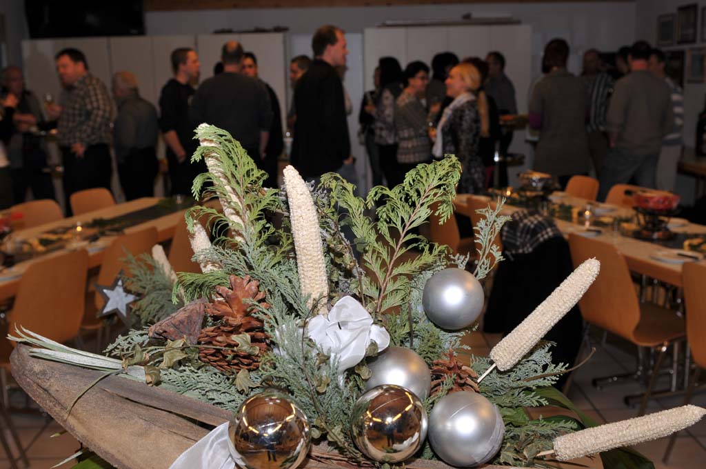 Gesteck im Vordergrund. Dahinter Personen am Apérölen.