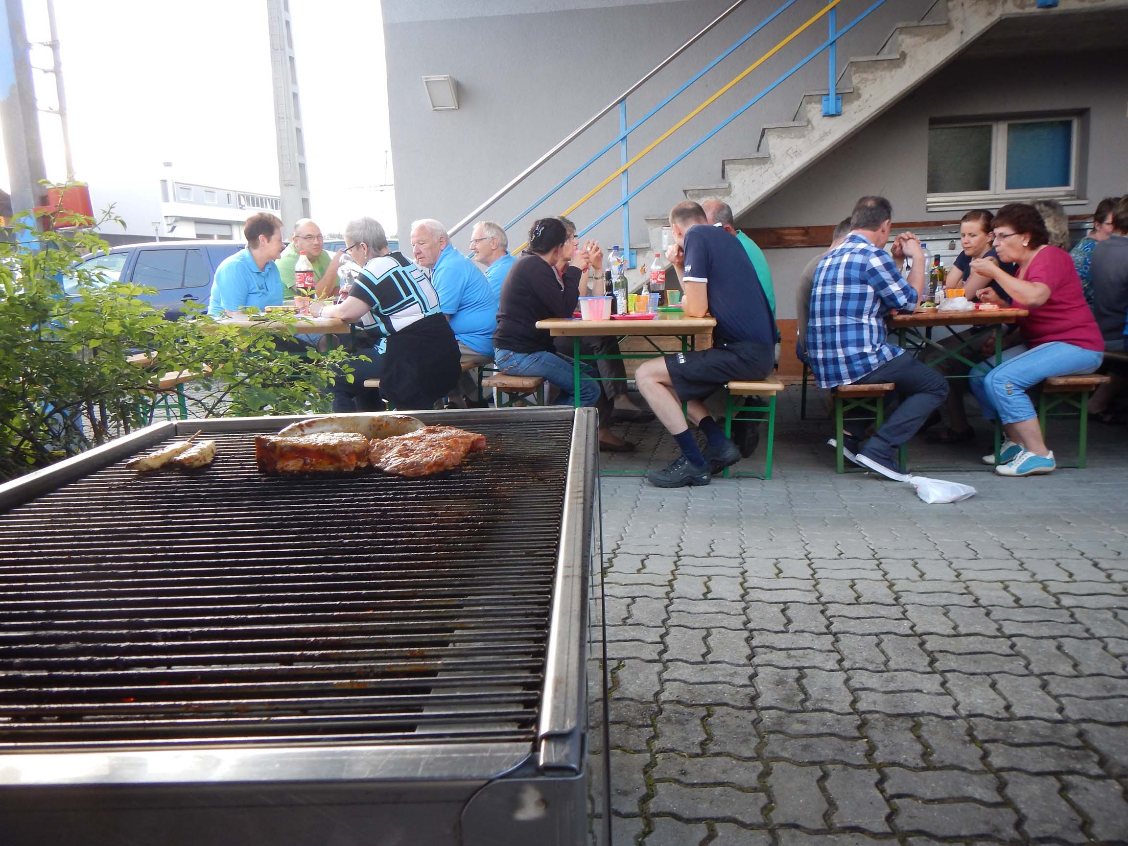 Im Vordergrund: Ein Grill mit Grillgut. Im Hintergrund: Festbänke mit Personen.