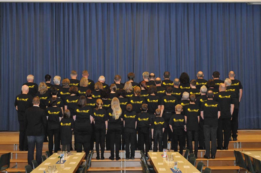 Die MG Uttigen auf der Treppe vor der Bühne. Den Rücken dem Fotografen zugewandt. So ist das Logo, MG Uttigen mit dahinter aufgehender Sonne, zu sehen.